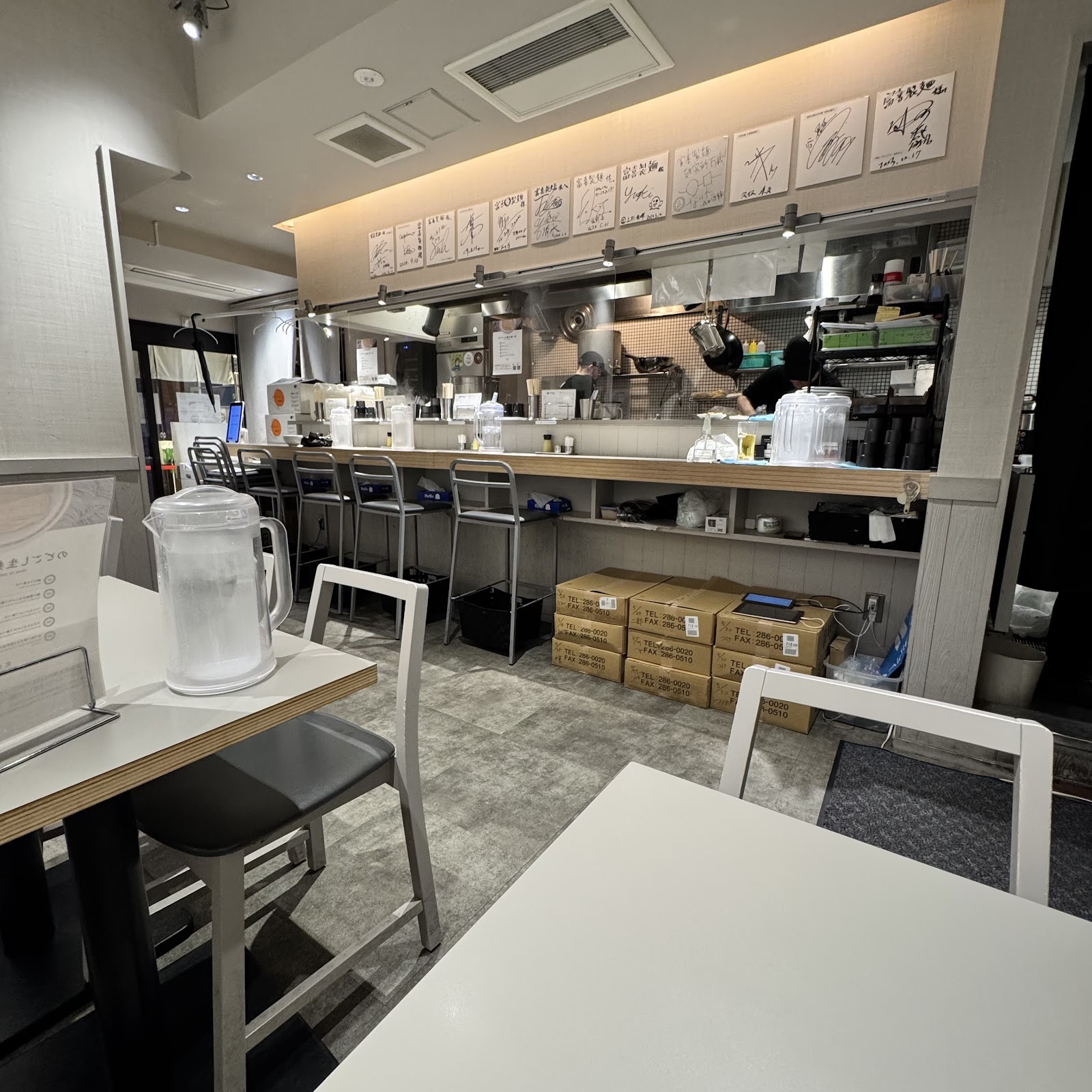 富喜製麺研究所の店内。手前に白いテーブルと椅子、奥にグレーのスツールが並ぶカウンターとオープンキッチン。厨房の上の壁に「富喜製麺」宛ての色紙が並ぶ。