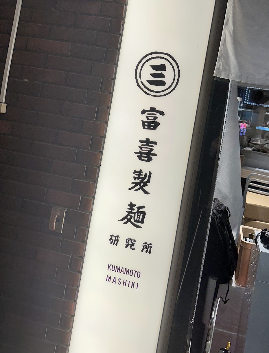 レンガの壁の横に立つ白い縦型看板「富喜製麺 研究所 KUMAMOTO MASHIKI」と、グレーの暖簾がかかった入口。暖簾の奥に厨房の明かりが見える。