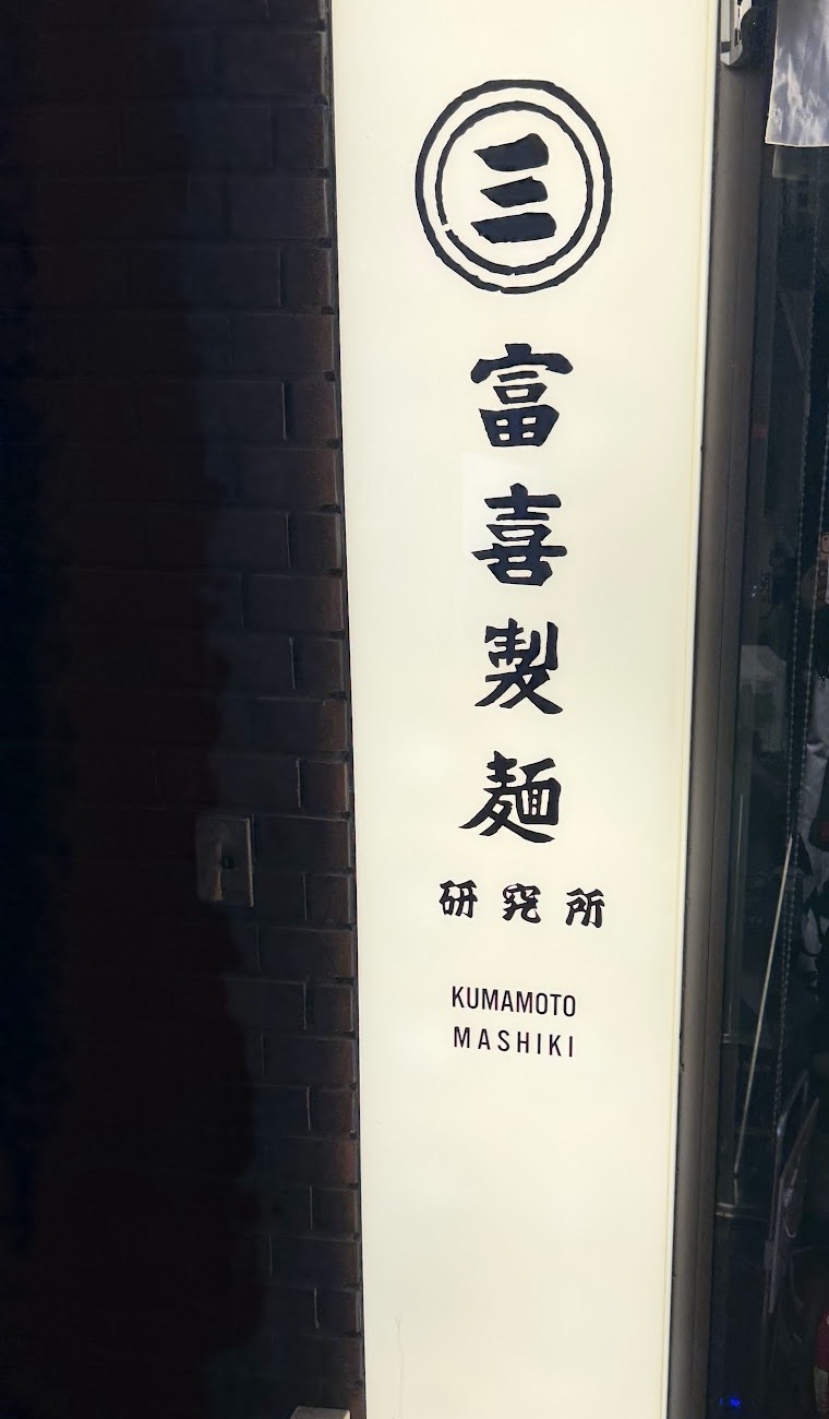 夜のレンガ壁に光る白い縦型看板。丸に三のマークと「富喜製麺 研究所」「KUMAMOTO MASHIKI」の文字。