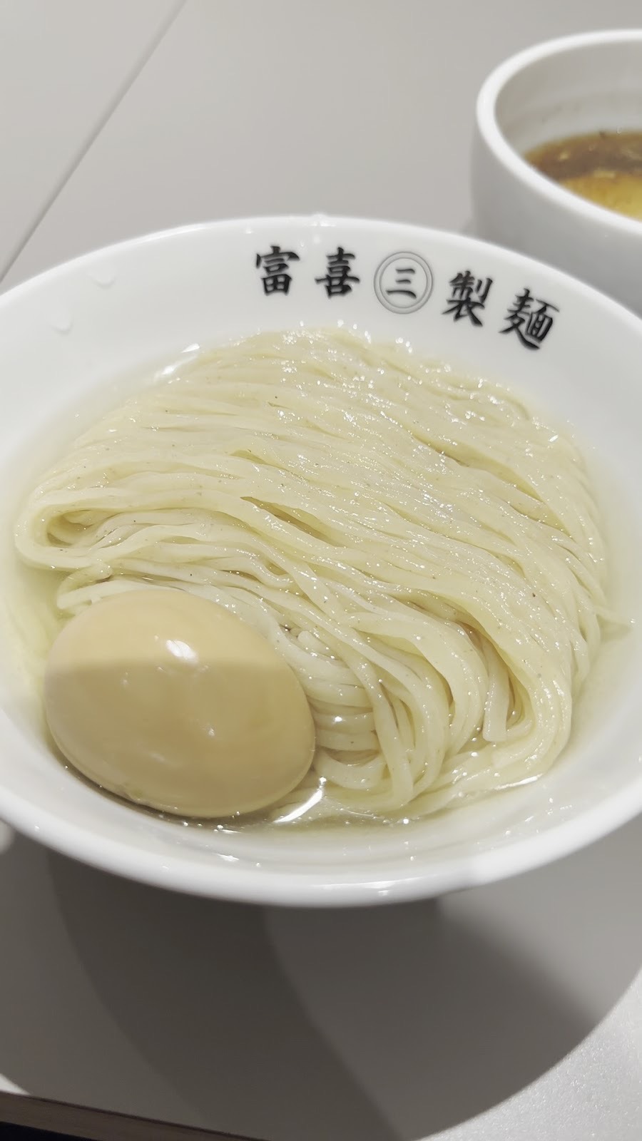 「富喜㊂製麺」と書かれた白い丼に、透き通った汁に浸かったつけ麺と味玉。奥につけ汁の器。