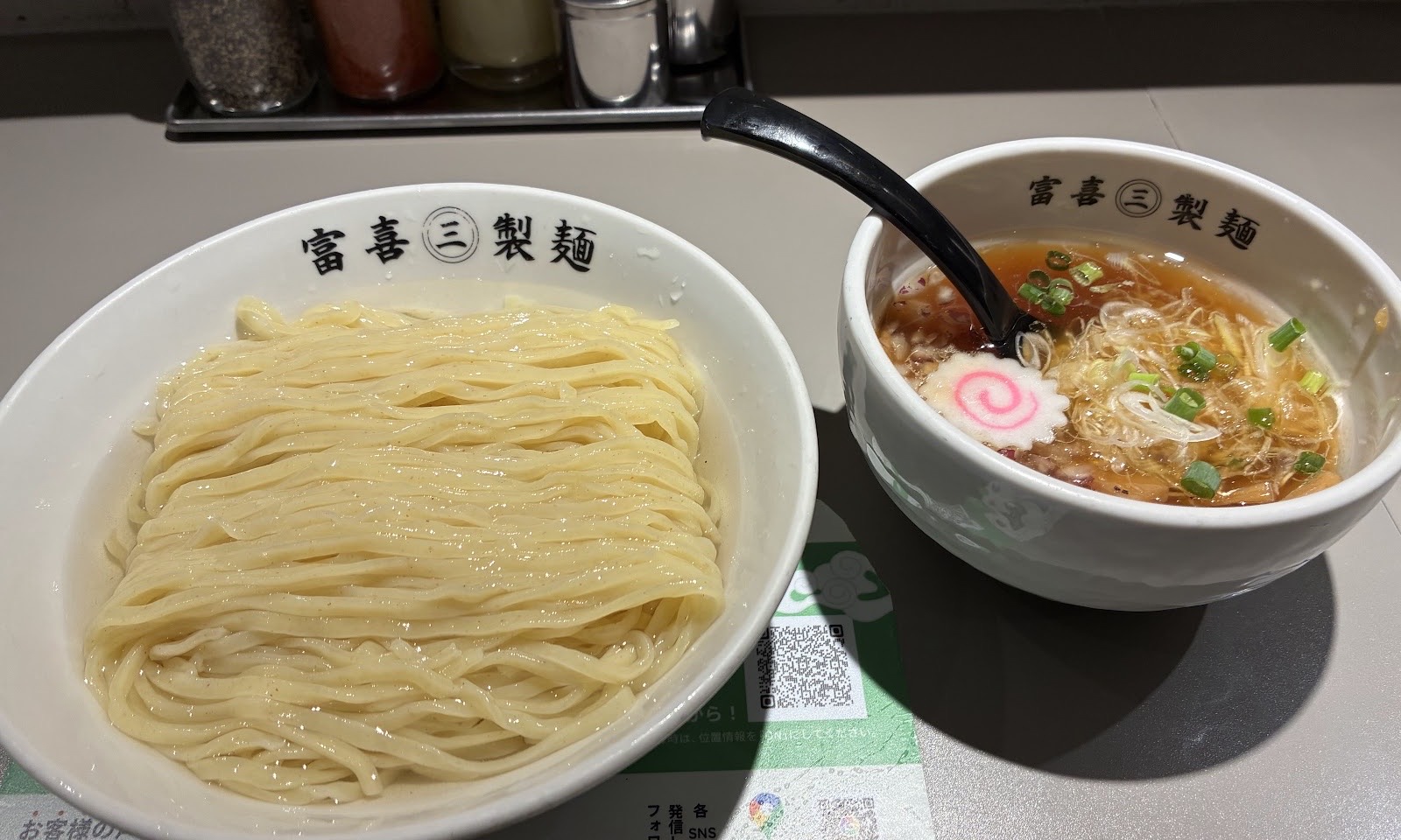 カウンターに並ぶつけ麺と、なるととねぎが浮かぶ醤油色のつけ汁。どちらも「富喜㊂製麺」の器。奥に調味料。