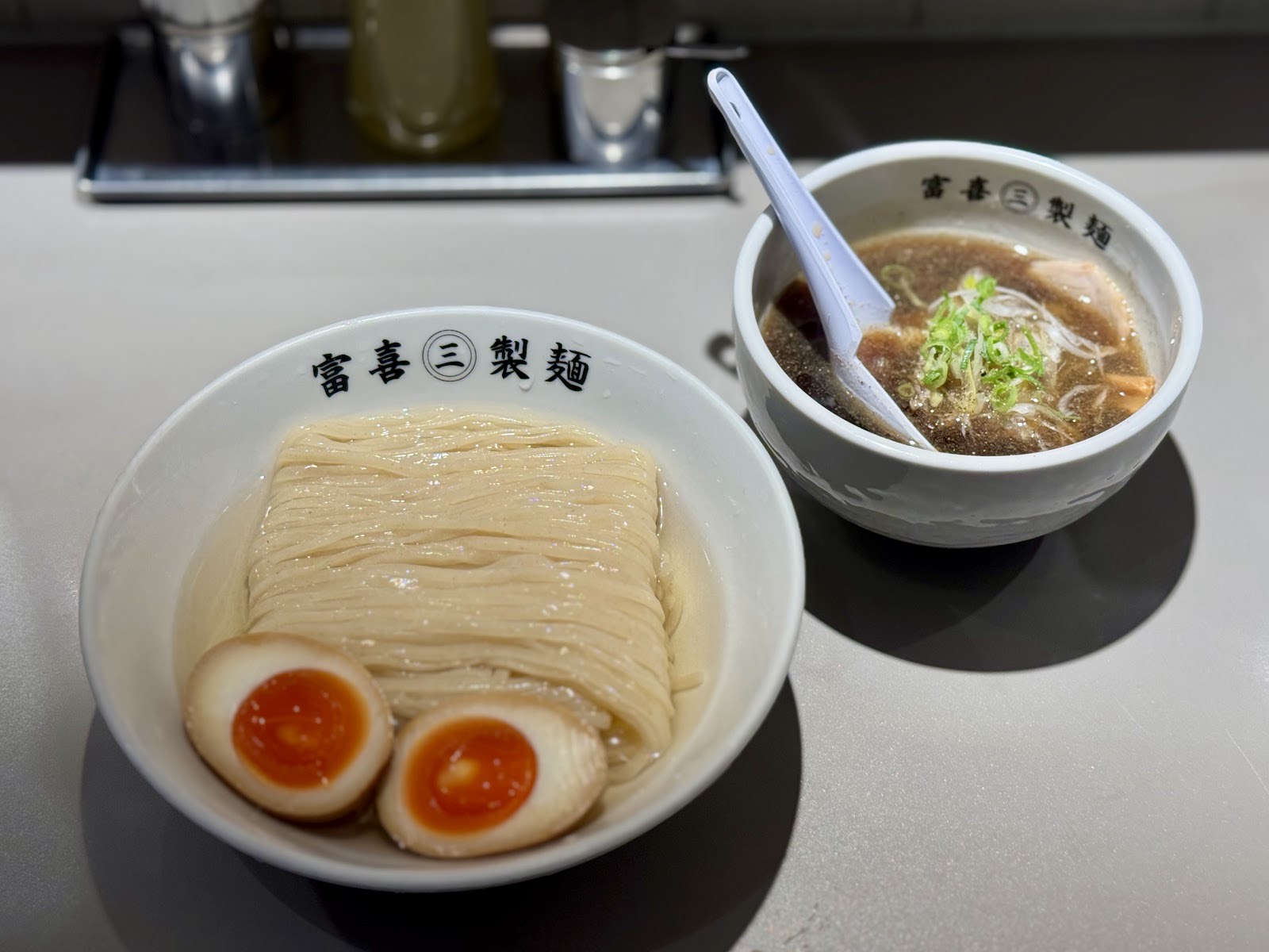 味玉がのったつけ麺と、ねぎの入ったつけ汁。「富喜㊂製麺」の器がカウンターに並ぶ。