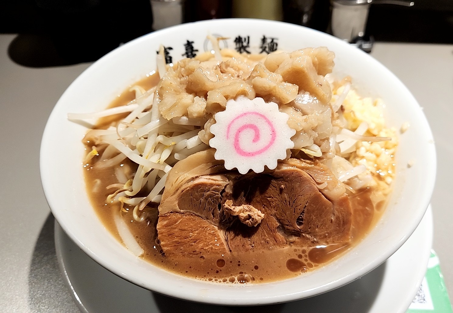 もやし、背脂、なると、ニンニク、厚切りの豚肉が山盛りのラーメン。「富喜㊂製麺」の丼。