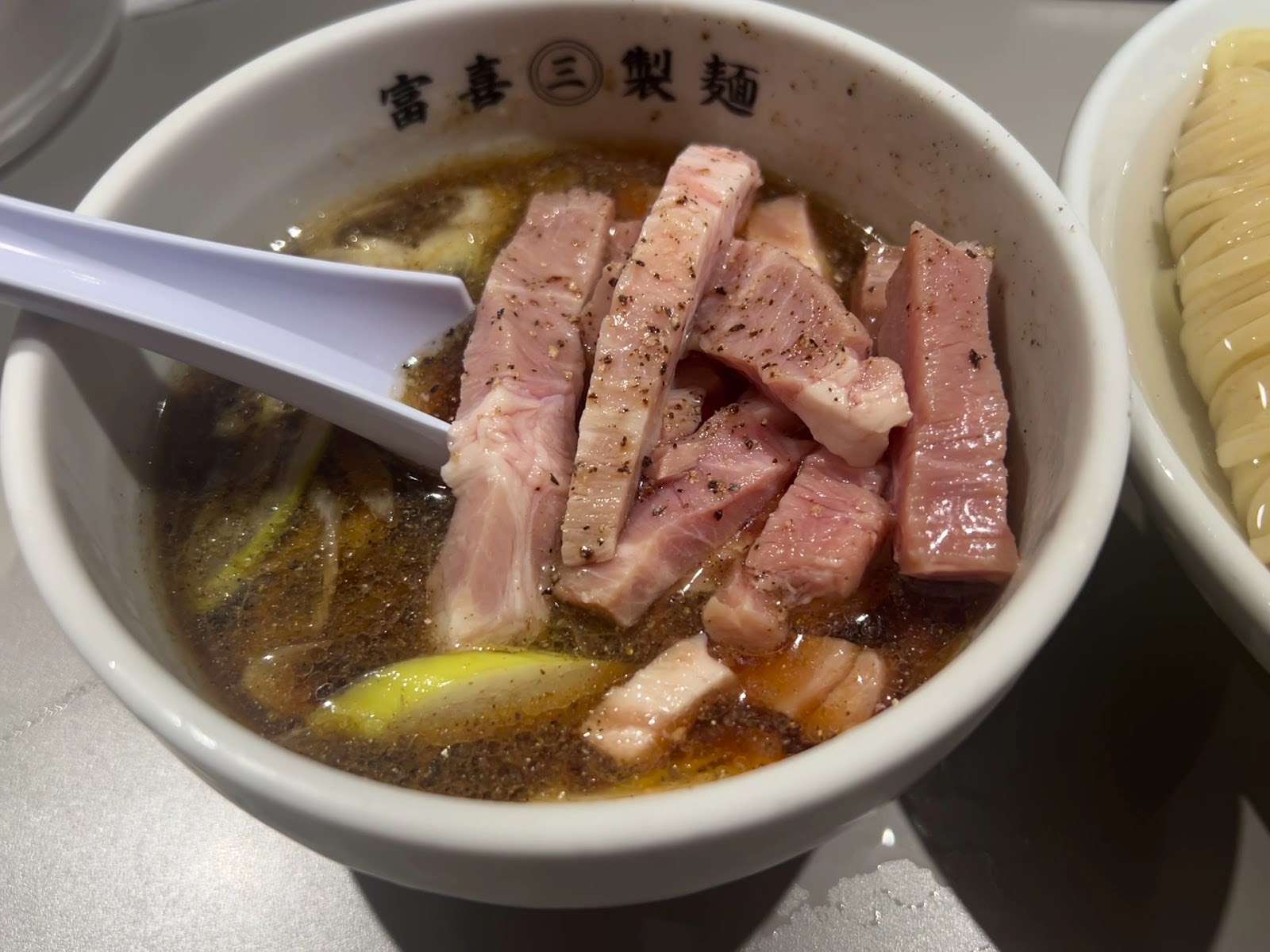 厚切りの豚肉とねぎが入り、黒胡椒が浮かぶつけ汁。「富喜㊂製麺」の器に白いレンゲ。
