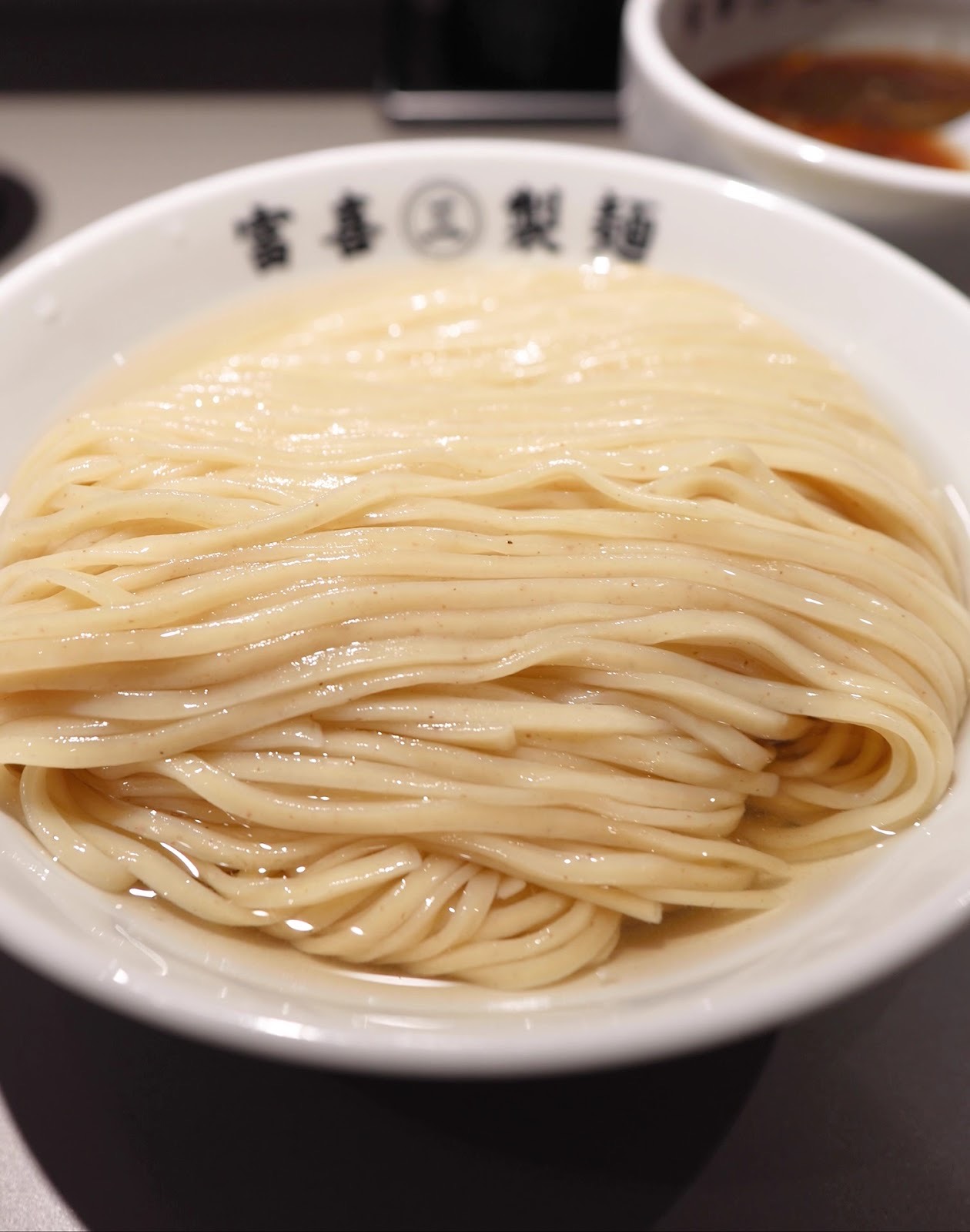 「富喜㊂製麺」の丼に整えて盛られたつけ麺の麺のアップ。奥に赤いつけ汁。