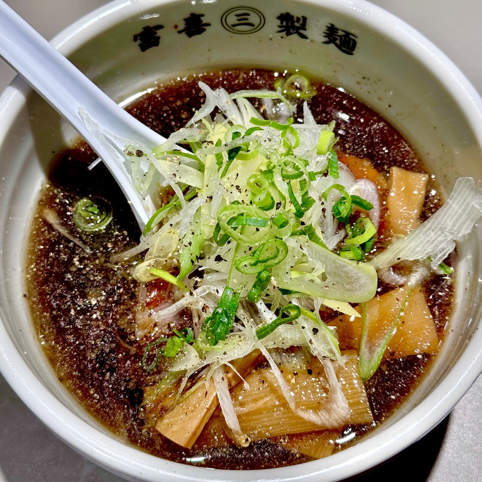 黒い醤油スープにメンマ、白髪ねぎ、青ねぎ、黒胡椒。「富喜㊂製麺」の丼に白いレンゲ。