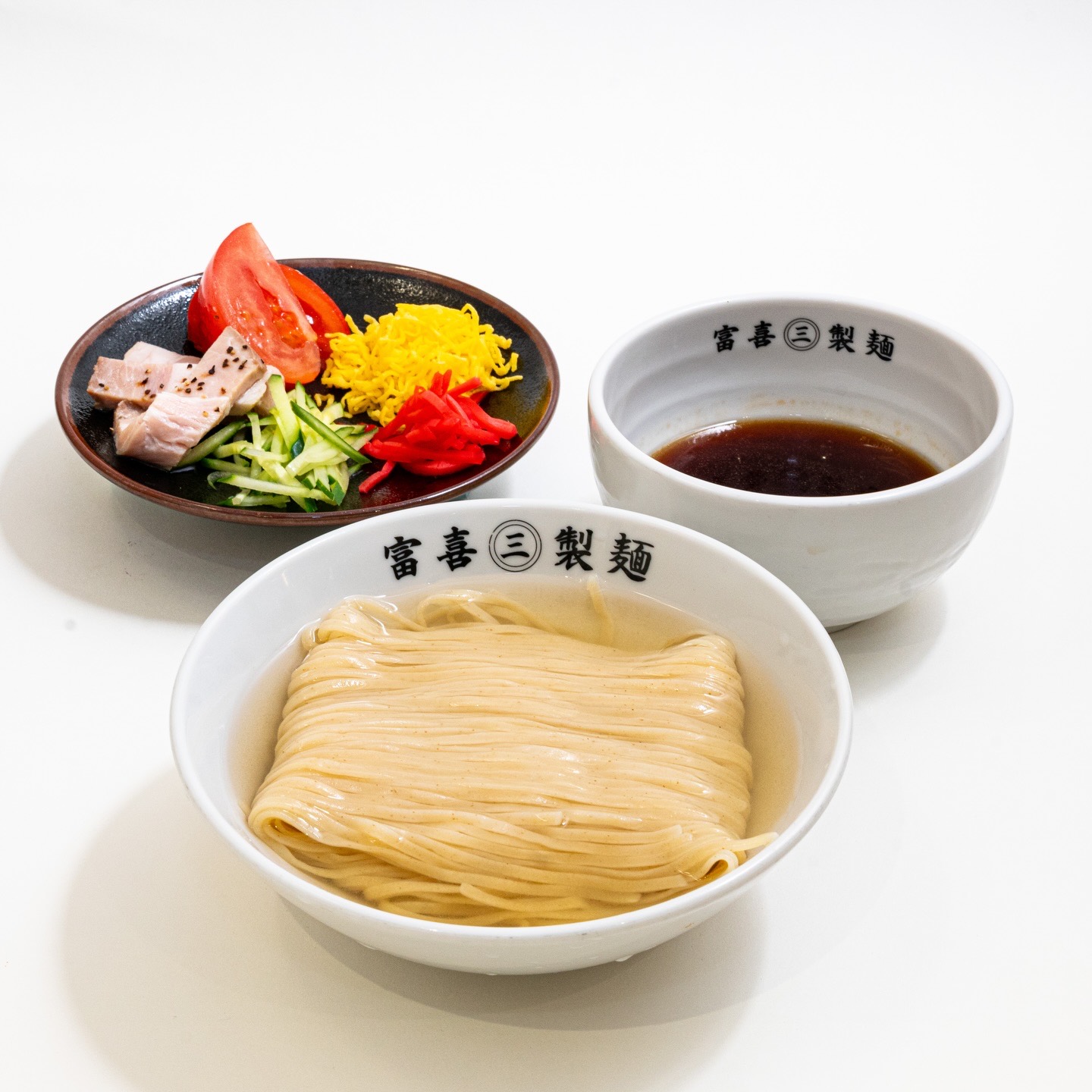 白い背景に並ぶ冷やし中華。トマト、錦糸卵、きゅうり、チャーシュー、紅しょうがの皿と、「富喜㊂製麺」の器の麺とたれ。