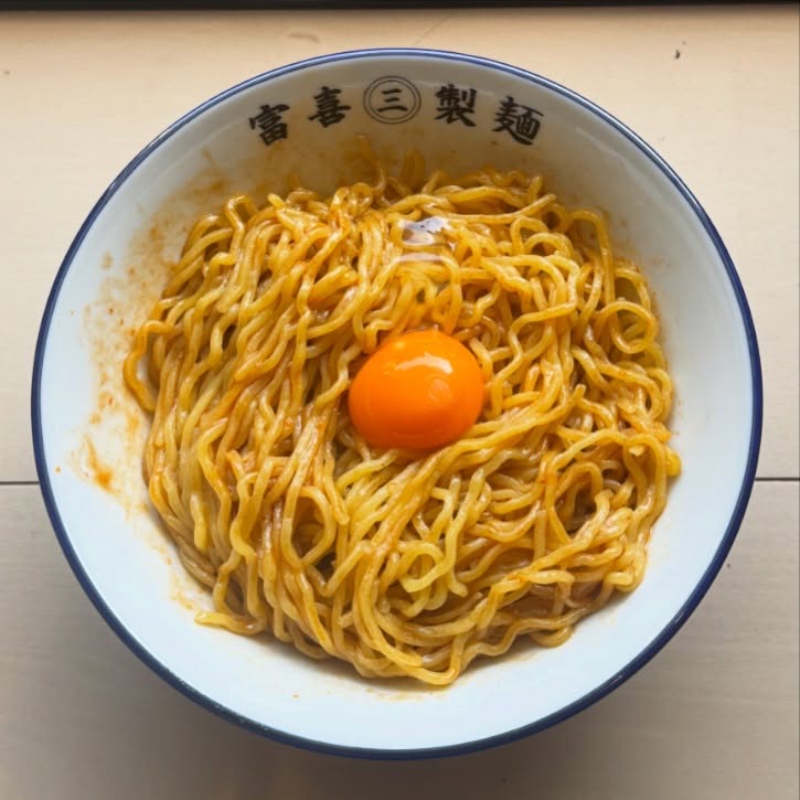 赤いタレをからめた黄色い麺の中央に卵黄。紺の縁の「富喜㊂製麺」の丼を上から。